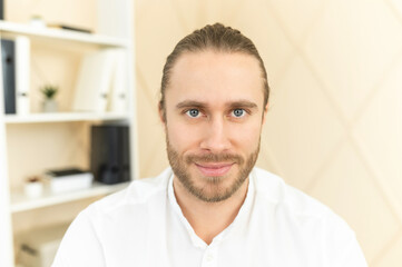 Handsome European man with light brown hair, gray eyes and beard looking at camera, smiling friendly, posing, confident, wearing white formal shirt, workplace, shelf with documents in the background