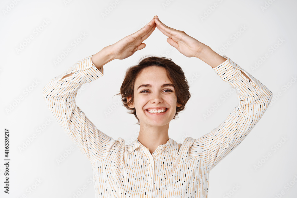 Wall mural Insurance and real estate concept. Candid woman smiling and showing house roof with hands above head, looking happy at camera, making rooftop gesture, white background