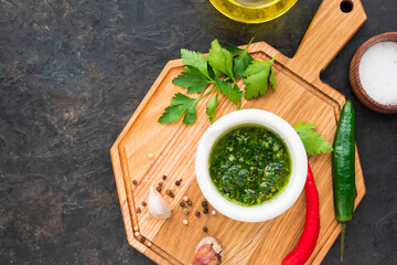 Chimichuri, Argentine green sauce for the steaks with fresh parsley, garlic, olive oil and oregano in a white mortar on a wooden board on a dark concrete background. Sauce recipes.