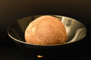 One ripe organic coconut on a black ceramic plate, close-up, isolated on black.