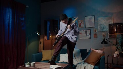 young musician man singing and playing guitar happy and crazy having fun jumping on home sofa couch listening to music and dancing, social distance, band guitarist on lockdown, streaming concert