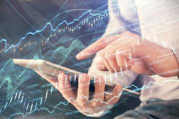Double exposure of man's hands holding and using a digital device and forex graph drawing....