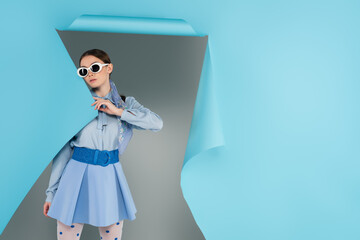 young fashionable woman in sunglasses near hole in blue paper on grey background