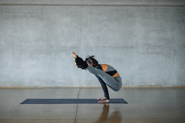 Young caucasian fit woman practicing yoga, doing Firefly pose, on the map on the floor, in front a gray wall, in sunny fitness studio. Instructor, professional pilates trainer. Healthy lifestyle.