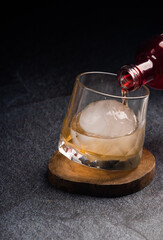 Pouring whiskey from bottle into glass with ice ball on table. Space for text