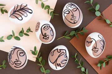 Happy Easter concept. Surreal faces on eggs and green branches of eucalyptus on brown background. Art und Online style. Top view Flat lay Copy space