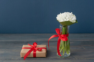 bouquet of white snowdrops in little transparent vase tired up with red ribbon, wrapped gift box is near on dark wooden table and blue background. Copyspace, greetings concept