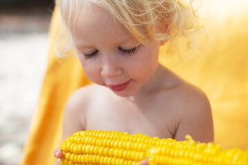 Blond and curly toddler girl eating corn on the beach. Yellow colors, summer mood