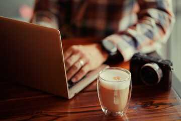 Traveler's coffee break. Cup of cappuccino on wooden table with a man typing on a laptop.