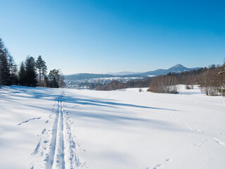 Winter landscape with view of village town Cvikov and ski run trail on snow-covered fields and snowy frozen forest and trees on sunny day, blue sky background