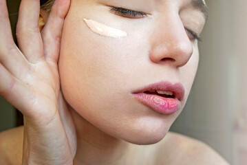 Young woman with bare shoulders and closed eyes applying foundation