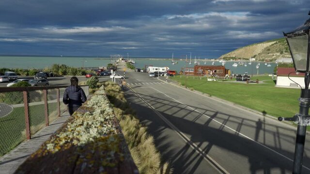 Brunette woman in a puffer jacket walks down the pavement towards Oamaru Harbour, New Zealand