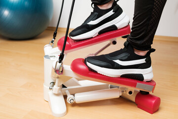 Fototapeta na wymiar Close up womans feet in sneakers training on the twist stepper with digital display and gum expanders at home during lockdown. 