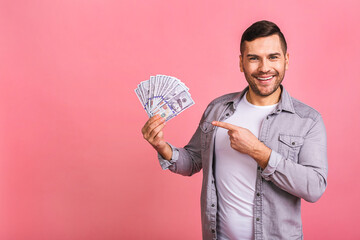 Happy winner! Young rich man in casual holding money dollar bills with surprise isolated over pink...