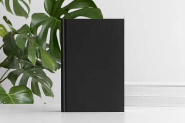 Black book mockup with workspace accessories on the white table and a monstera plant.