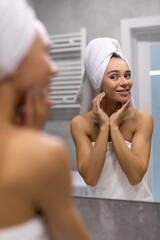 Beautiful young woman in bath towel is touching her face and smiling while looking into the mirror...