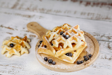 On a light background, Viennese waffles are stacked on a round board, one on one. Golden waffles are decorated with black currants.