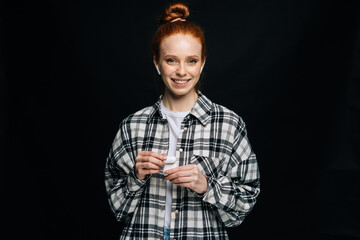 Smiling red-haired young woman wearing wireless earphones looking at camera on isolated black background. Pretty redhead lady model emotionally showing facial expressions, copy space.