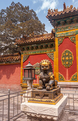 Beijing, China - April 27, 2010: Forbidden City. Closeup of small brown-golden lion statue on white pedestal with red-orange-greeen artwork in back. Blue cloudscape and green foliage.