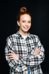 Happy red-haired young woman wearing wireless earphones looking at camera with cross hands on isolated black background. Pretty redhead lady model emotionally showing facial expressions.