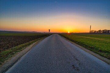 Straße in Bayern, Deutschland mit Blick auf den Sonnenuntergang am Horizont, welcher den Himmel in prächtigen Farben erstrahlen lässt.