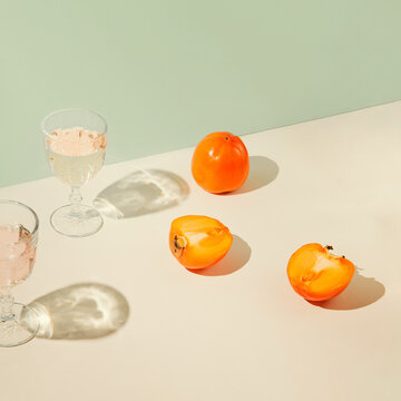 Persimmon fruits and glasses of water on geometric pastel background with sunlit. Summer drinks and refreshment concept.  Minimal style composition