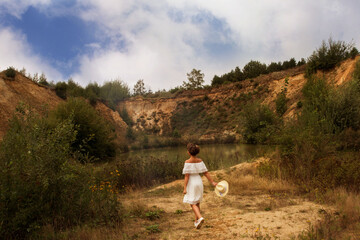 Junge Frau im weißen Kleid läuft zum See  und hält den weißen Hut an der Hand