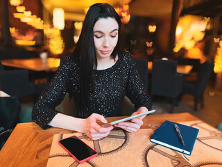 A girl with dark hair is working in a cafe with a tablet, a phone and a notebook are nearby....