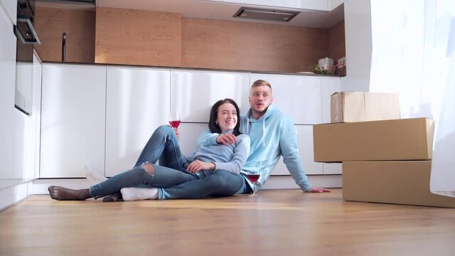 young happy married couple man and woman sitting on the floor and celebrating moving to a new apartment or home. They drink wine and plan the interior near the cardboard boxes. dream of modern design
