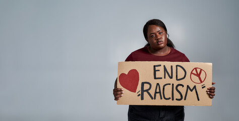 Tired plus size young african american woman in casual clothes looking at camera, holding End Racism banner in front of her, posing isolated over gray background