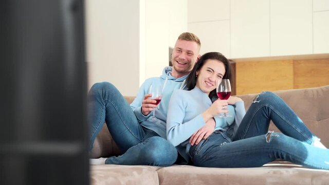 young cheerful couple watching funny movie on tv at home. A man and a woman or a boyfriend and a girlfriend are sitting on the couch with a glass of wine watching a TV series and laughing.