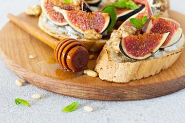 Crispy bruschetta with soft ricotta, ripe figs, walnuts and pine nuts, mint and honey on a light background. Figs fruit toast on a wooden board with honey and walnut.
