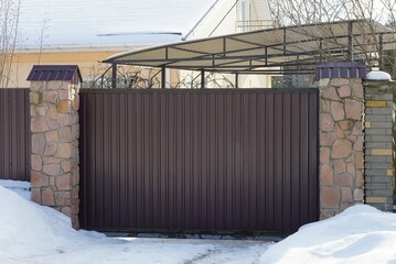 closed brown metal gates and part of the wall of a fence made of stones in white snowdrifts on a winter street