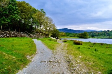 Fototapeta na wymiar Pretty Shot Down Lake District Pathway of Distant Hikers, Ancient Stone Wall, Mountains and Lake to the Side, Grasmere