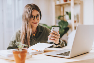 Freelance woman in glasses with mobile phone typing at laptop and working from home office with plants. Happy girl on workplace at the desk. Distance learning online education and work.
