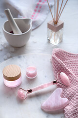 Crystal rose quartz facial roller and Gua sha stone for face massage. Pink skin care products and oils. Off white marble and textile background, natural light.