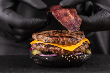 Close-up, making grilled burger. Chef hands in black gloves are puts slice of bacon in burger with...