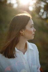 Fototapeta premium young woman in white linen clothes stands and looks to the side against the background of forest, sand and greenery at sunset