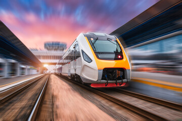 Fototapeta na wymiar High speed train in motion on the railway station at sunset. Fast moving modern passenger train on the railway platform. Railroad with motion blur effect. Commercial transportation. Front view