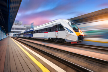 Naklejka premium High speed train in motion on the railway station at sunset. Modern intercity passenger train with motion blur effect on the railway platform. Industrial. Railroad in Europe. Commercial transportation