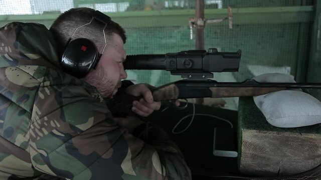 Male shooter hunter takes aim looking through the sight. Shooting training in the shooting range on the meshes. A man in camouflage clothes and headphones is ready to shoot at a target
