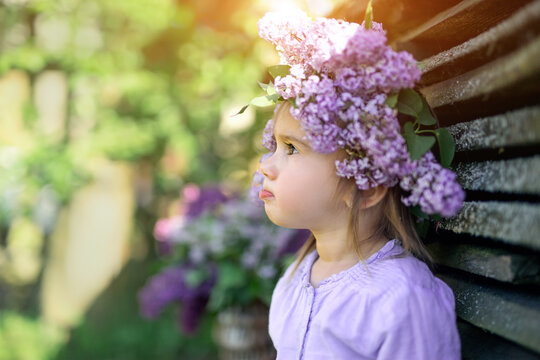 A girl with a wreath of lilac on her head looks away, pursing her lip. The pensive and offended look of a child.  May evening sunset. The child looks to the side.