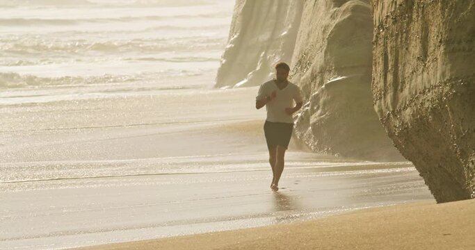 Footage of a runner during workout among beautiful natural landscape. Male jogger runs across sandy beach by the cliffs. High quality 4k footage