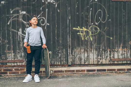 Teenager skateboarder boy standing beside a wooden grunge graffiti wall with skateboard and Water bottle flask. Youth generation Freetime spending and active people concept image.