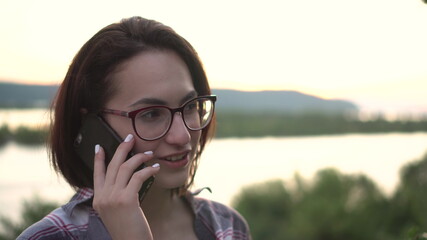 A young woman stands against the background of the river and mountains and speaks on the phone.