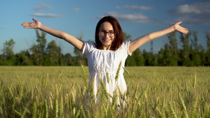 A young woman in a white dress jumps out of a green wheat field. The girl suddenly appears and smiles.