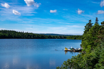 Three Adirondack chairs blue, green and yellow in colour, on a wooden floating boating dock near a shore covered in green lush trees. The calm smooth pond is deep blue. The sky is blue with clouds.