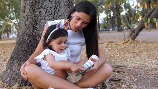 Madre e hija con telefono celular en parque navegando sentadas en árbol