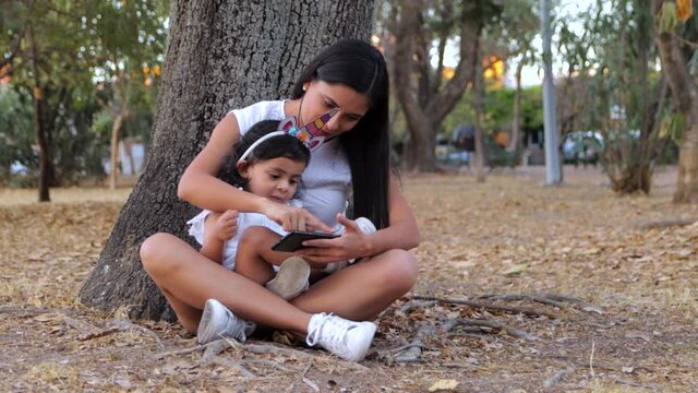Madre e hija navegando en tel&eacute;fono m&oacute;vil sentadas al pie de un &aacute;rbol en parque