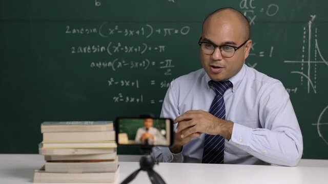Indian young teacher man sitting teaching online video conference live stream by smartphone. Asian teacher teaching mathematics class webinar online for students learning.